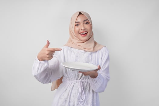 Smiling young Asian Muslim woman in a veil hijab pointing to an empty plate with copy space over it, presenting food menu for iftar. Ramadan concept photo