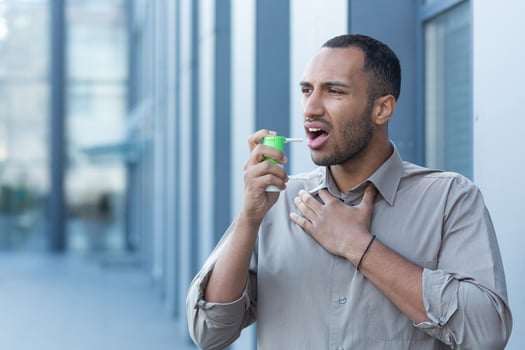 A young Latin American man clears his throat with an inhaler and medication. He stands outside photo
