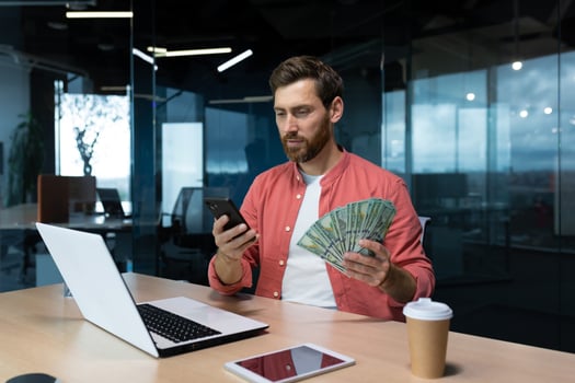 A young businessman, freelancer, banker sits in the office at a table with a laptop, holds cash money in his hands, uses a telephone. photo