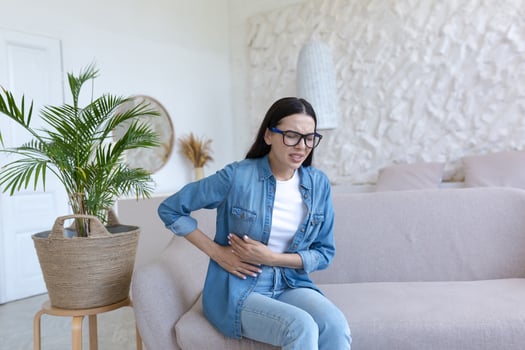 A young woman is sitting on the sofa at home, grimacing and holding her stomach. Suffers from severe abdominal pain, menstrual pain, pregnancy problems, miscarriage, poisoning, spasms. photo