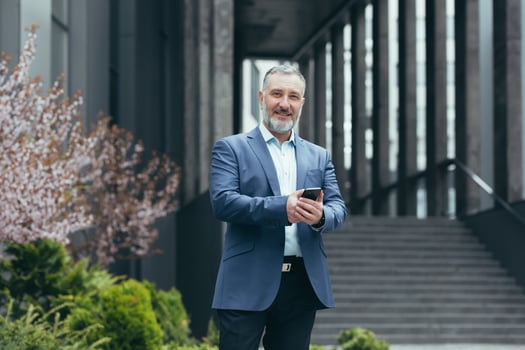 Portrait of successful senior gray-haired banker outside office building, man with phone smiling and looking at camera, businessman happy in business suit photo