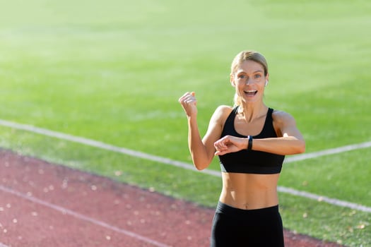 Sporty young woman athlete standing in the stadium wearing headphones and smiling at the camera, checking the time on the fitness bracelet, showing a victory gesture with her hand. photo