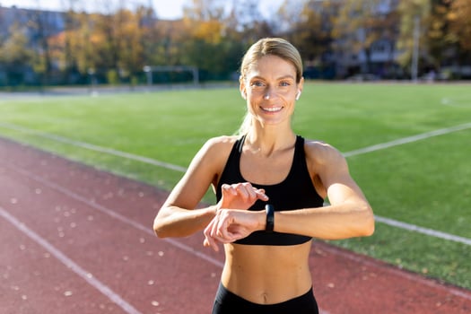 Portrait of a sporty young woman standing at the stadium wearing headphones and smiling at the camera, checking the time on a fitness bracelet, looking at the time of running, classes. photo
