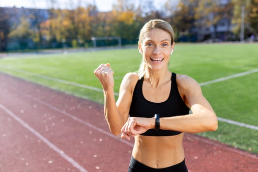A sporty young woman standing in a stadium wearing headphones and smiling at the camera, checking the time on a fitness bracelet, looking at the time of a run, showing a victory gesture with her hand. photo