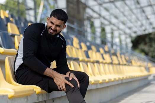 A male athlete in workout attire experiencing knee pain while sitting on yellow stadium seats, highlighting sports injury and fitness concepts. photo