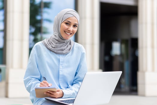 A young Muslim woman focused on studying outside, taking notes beside her laptop at a university campus. photo