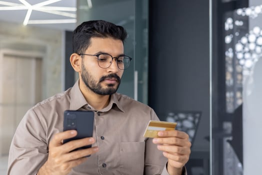 Focused young businessman in glasses using a smartphone and holding a credit card, possibly making an online purchase or managing finances in a modern office space. photo