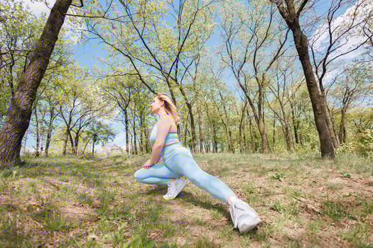 The girl extends her leg out to the side thereby stretching the leg muscles Beautiful blonde Caucasian woman in blue tight tracksuit. Blonde girl at an outdoor training session photo