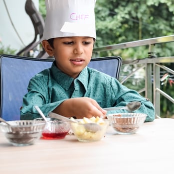 Cute Indian chef boy preparing sundae dish as a part of non fire cooking which includes vanilla ice cream, brownie, coco powder, freshly chopped fruits and strawberry syrup. Little kid preparing food photo