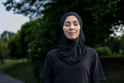 A young woman wearing a hijab stands outdoors with her eyes closed, exuding a sense of peace and serenity surrounded by nature. photo