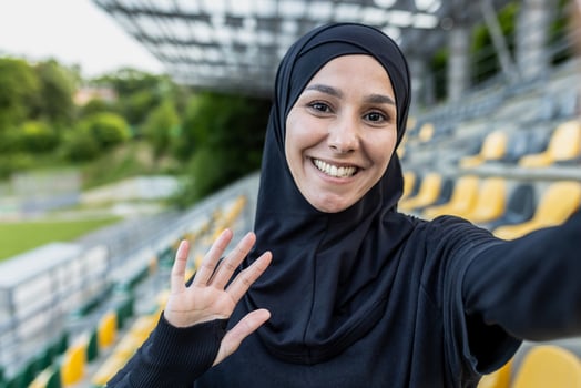 Cheerful Muslim woman in hijab taking a selfie at a sports stadium, waving and smiling at the camera. photo