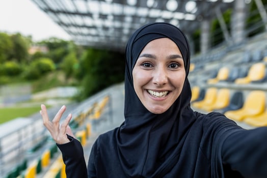 A cheerful Muslim woman in a hijab captures a selfie moment with a wide smile at a sunny sports stadium with yellow seats. photo