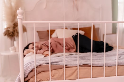 A woman is laying on a bed with a white metal frame. She is wearing a pink shirt and is resting her head on her hand. The bed is covered in a white blanket and pillows. photo