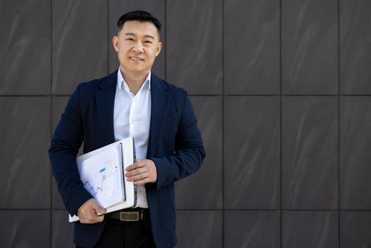 Portrait of a smiling young Asian man in a business suit standing against the wall of an office building, holding documents and looking confidently at the camera. photo