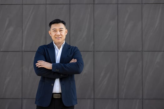 Portrait of young successful Asian businessman, lawyer in suit standing outside against wall background, arms crossed on chest and smiling confidently at camera. photo