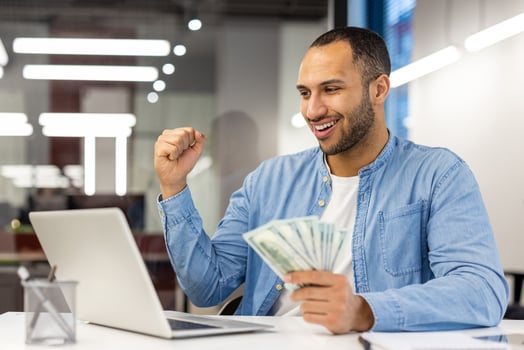 A happy businessman at a desk with a laptop, cheerfully celebrating financial success while holding cash in hand. photo