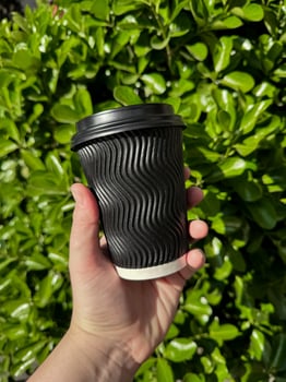 Hand holding black corrugated cardboard coffee cup with lid against background of lush green leafy bush. Urban eco friendly lifestyle. Blank space for sticker or label. photo