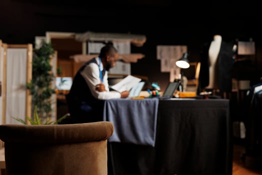Hip level shot of cozy tailoring studio with blurry african american dressmaker in the background studying sartorial designs. Couturer inspecting sketches of upcoming fashion collection photo