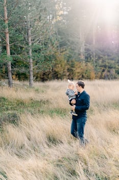 Dad with a little girl in his arms walks through the tall grass at the edge of the forest photo
