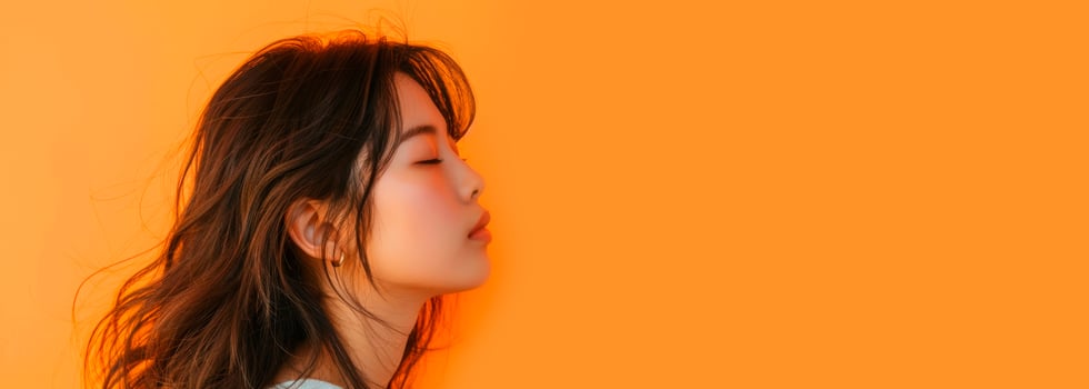 A woman with black hair stands in front of an orange wall, eyes closed, smiling photo