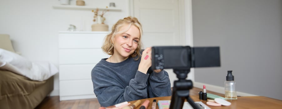Image of young stylish woman, blogger recording a beauty lifestyle of her picking best lipstick, showing lip balm swatches on her skin, sitting in front of digital camera in empty room photo