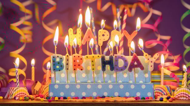 Lit candles of different colors in the shape of letters forming the phrase HAPPY BIRTHDAY, against a purple out-of-focus background photo