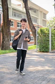 A happy Asian male college student is using his smartphone while walking in the campus park. photo