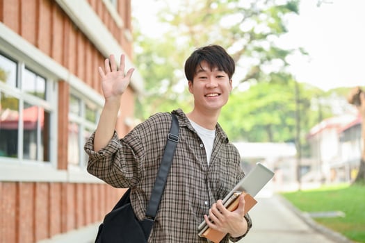 A friendly Asian male student stands outdoors on campus, waving his hand and smiling at the camera. photo