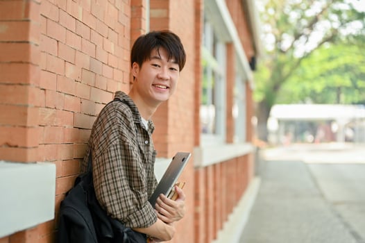 A smiling young Asian male college student stands by a brick wall on campus, carrying a backpack. photo