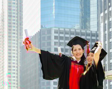 Two happiness graduated female in black graduation gowns holding diploma and hugging each other on graduation ceremony, standing outdoors with city buildings background photo