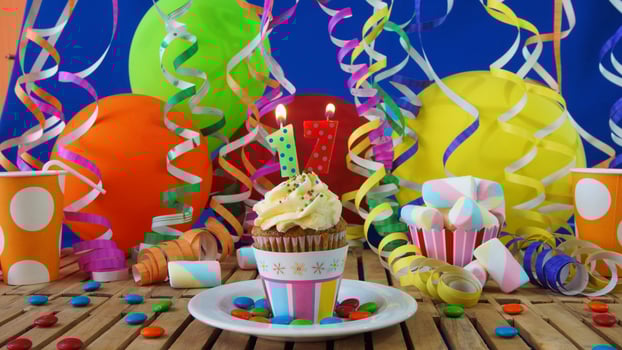 Birthday 17 cupcake with candles burning on rustic wooden table with background of colorful balloons, plastic cups and candies with blue wall in the background photo
