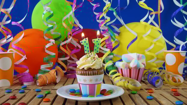 Birthday 11 cupcake with candles burning on rustic wooden table with background of colorful balloons, plastic cups and candies with blue wall in the background photo