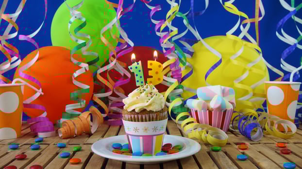 Birthday 13 cupcake with candles burning on rustic wooden table with background of colorful balloons, plastic cups and candies with blue wall in the background photo