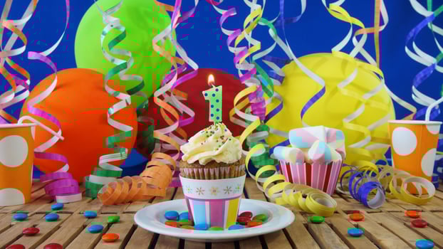 Birthday 1 cupcake with candles burning on rustic wooden table with background of colorful balloons, plastic cups and candies with blue wall in the background photo