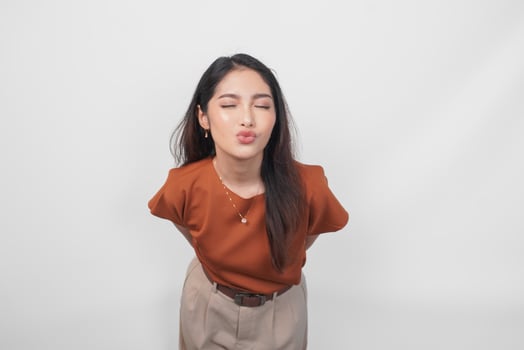 Cute Asian woman in brown shirt sending blowing kiss with pout lips looking at camera isolated on white background. photo