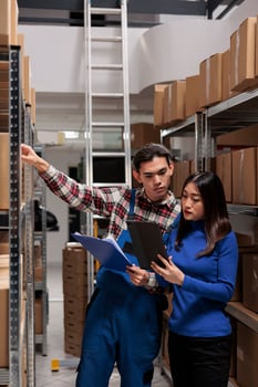 Warehouse employees managing inventory tracking with goods checklist. Storehouse asian man and woman workers picking order while standing in storage room aisle with shelves full of cardboard boxes photo