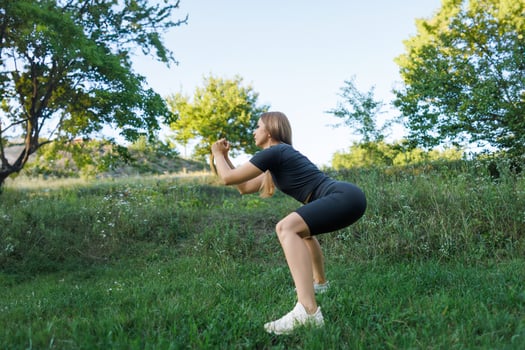 The girl performs athletic squats with her arms folded in front of her. Athletic girl in a tight uniform working outdoors in the park. photo