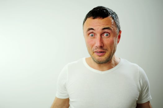 attractive unshaven man astonished amazed in shock surprise face expression and shock emotion Blue eyes black hair with gray White t-shirt in the studio photo