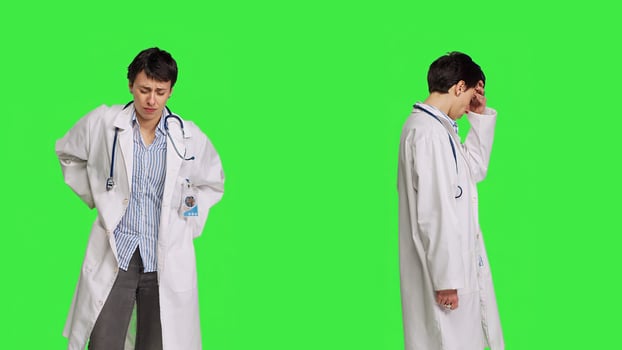 Medic in white coat suffering from migraine and lower back pain, feeling overworked and stressed at hospital. Unwell doctor with headache and spine discomfort against greenscreen backdrop. Camera B. photo