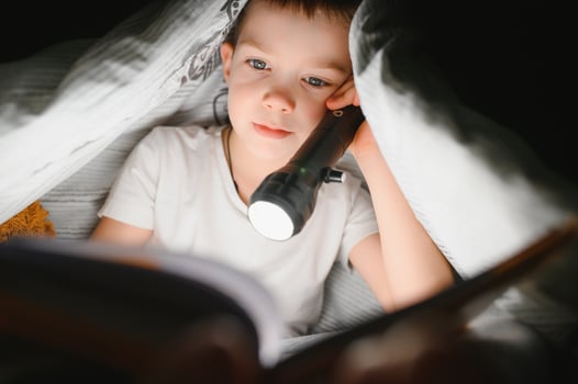 Boy with flashlight reading book under blanket at home. photo