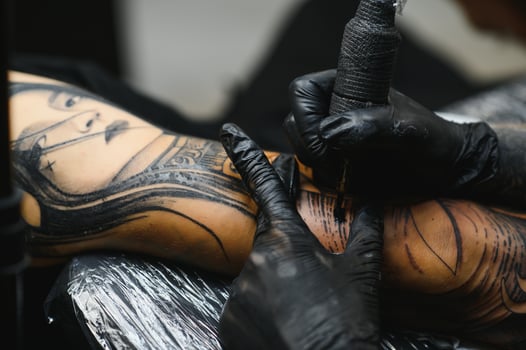 Cropped close up of a bearded tattoo artist working at his studio tattooing sleeve on the arm of his male client. Man getting tattooed by professional tattooist photo
