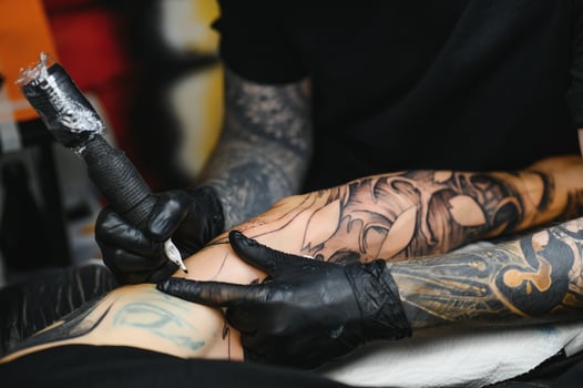 Cropped close up of a bearded tattoo artist working at his studio tattooing sleeve on the arm of his male client. Man getting tattooed by professional tattooist photo