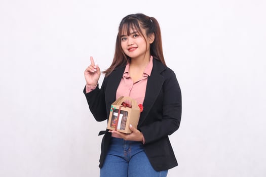 Beautiful Asian girl in her 20s smiles excitedly pointing up at Christmas gift in the form of hamper in hand on white background for advertising materials, banners, billboards photo