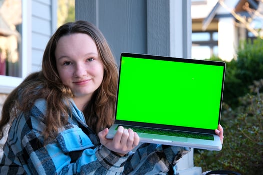 Portrait studio shot of young hipster female model in casual street wears sunglasses sit on floor holding pointing green blank empty screen laptop computer for advertising photo