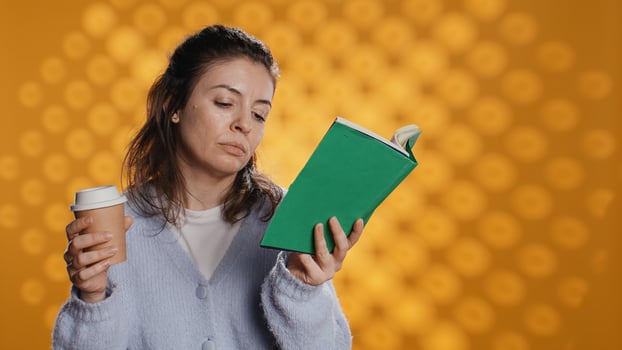 Tired woman reading book, holding cup of coffee, isolated over studio background. Exhausted bookworm enjoying novel and caffeine beverage, lacking energy, struggling to stay awake, camera B photo