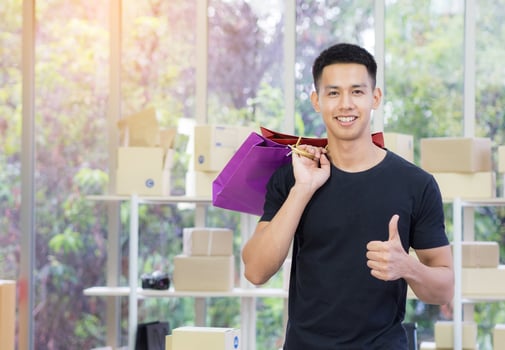 Cheerful young man holding colorful shopping bags, giving thumb up, and looking at the camera while standing in the office with sunlight. Concept of business and e-commerce photo
