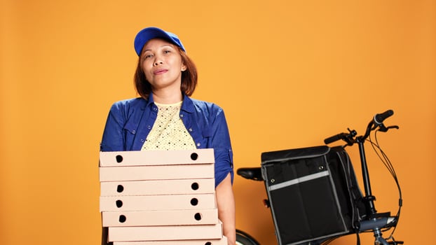 Impatient asian worker waiting for customer to answer door and pick up food. Portrait of bored courier isolated over orange studio background carrying pile of pizza boxes photo