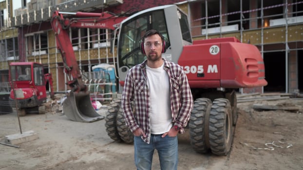 Portrait of successful builder, worker, contractor wearing hard hat and protective earphones, standing on work site with crossed arms confidently. Heavy construction equipment in background. photo