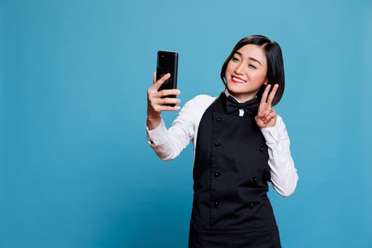 Smiling asian waitress holding smartphone, looking at front camera and showing peace symbol with fingers. Cheerful woman receptionist posing while taking selfie on mobile phone photo