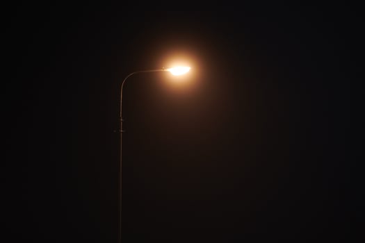 One night lamppost shines with faint mysterious yellow light through evening fog, copy space photo